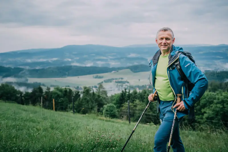 Andreas Kohlbacher mit Wanderstöcken auf einer Wiese über den Hügeln der Weststeiermark